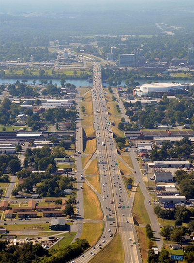 Aerial photo of the I-30 Corridor shows on- and off-ramps connecting to parallel, local access roads. Development and stands of trees flank the corridor on both sides. In the distance, the I-30 bridge crosses the Arkansas River into Downtown Little Rock.