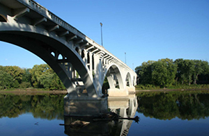 Photograph of a bridge over a calm river. Viewpoint is from river level, looking up at the underside of the bridge with clear blue sky.