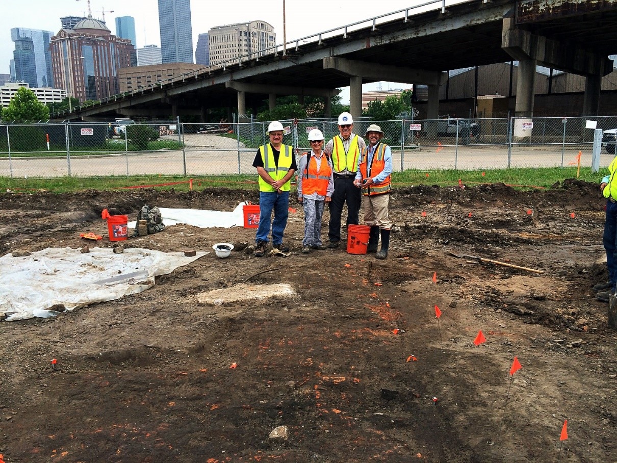 A group of four people stand on a construction site with a highway and cityscape in the background.