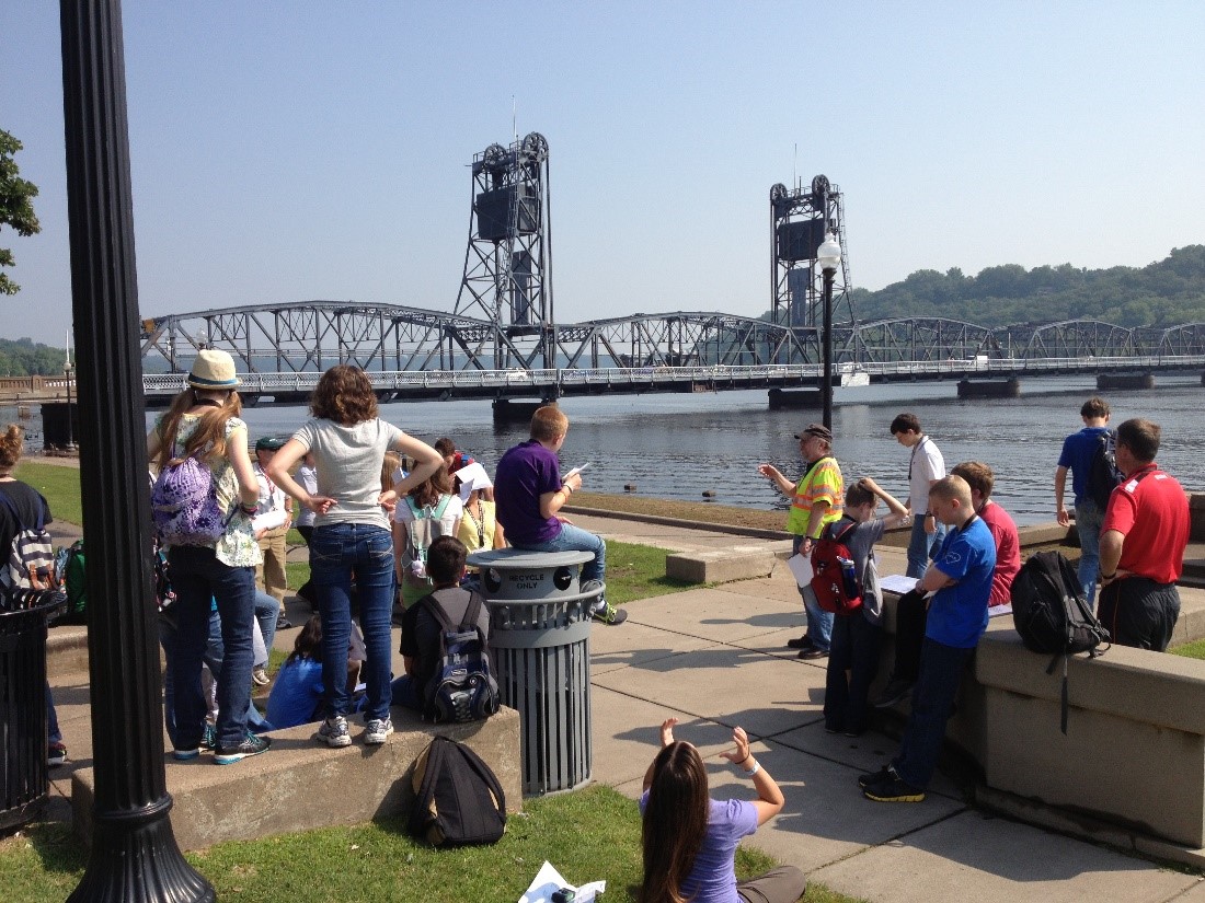 Several people looking out across a body of water at a historic lift bridge that stretches the width of the water.