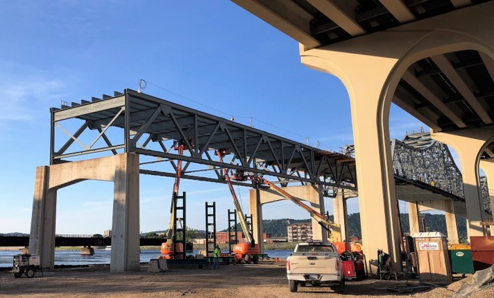 A photograph of a truss bridge being constructed that runs parallel to an existing bridge on the right side of the image.