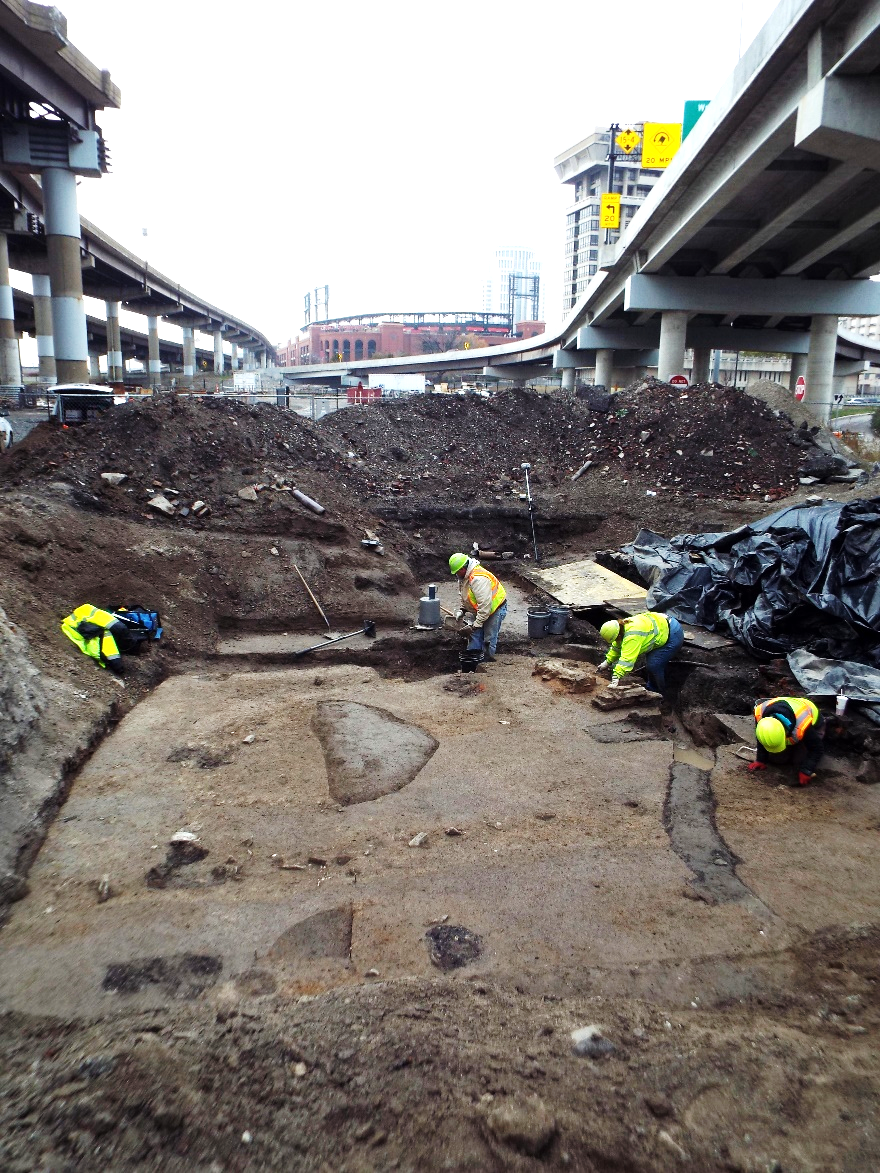 A photograph of four people in hard hats and safety vests investigating an archeological site.