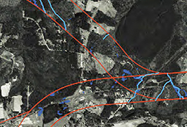 A black and white aerial photograph. Wetlands and streams are delineated in dark and light blue, respectively. Red lines indicate potential alignments for a proposed transportation facility.