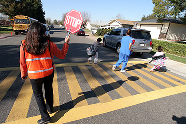 Photograph of a crossing guard holding up a STOP sign in the middle of a crosswalk as a woman and two school age children cross
