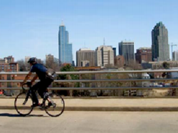 photograph of a bicyclist traveling along a raised roadway with a decorative guardrail; large buildings are in the background