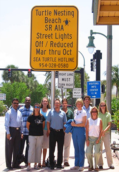 Photograph of the 2008 Broward County, Florida Sea Turtle Friendly Lighting Team standing below a large 'Turtle Nesting Beach | SR A1A | Street Lights Off/Reduced | Mar thru Oct | Sea Turtle Hotline | 954-328-0580' caution sign on a sunny day