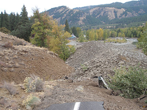 Photograph of a section of State Route 410, showing the road damage resulting from a landslide. The road is covered with large mounds of dirt and rocks and uprooted plants and trees.