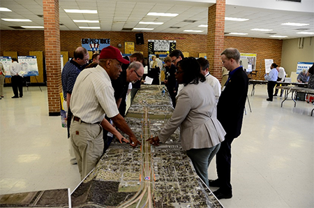 Photo of participants and facilitators at a public meeting stand around a long table with large-format aerial maps of the I-30 corridor discussing and pointing to features on the map.