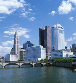 Photograph of a cluster of modern buildings clustered along a river. A bridge crosses the river in front of the buildings.