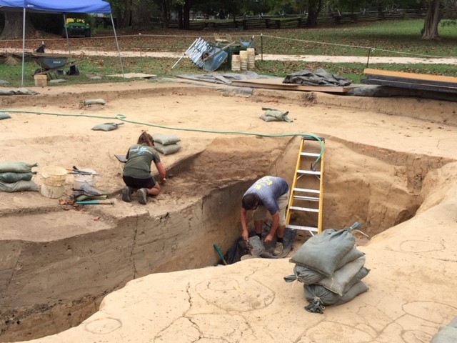 A photograph of a dig site with two people working on excavating the site. There are tools, a tent, and ropes enclosing the site.