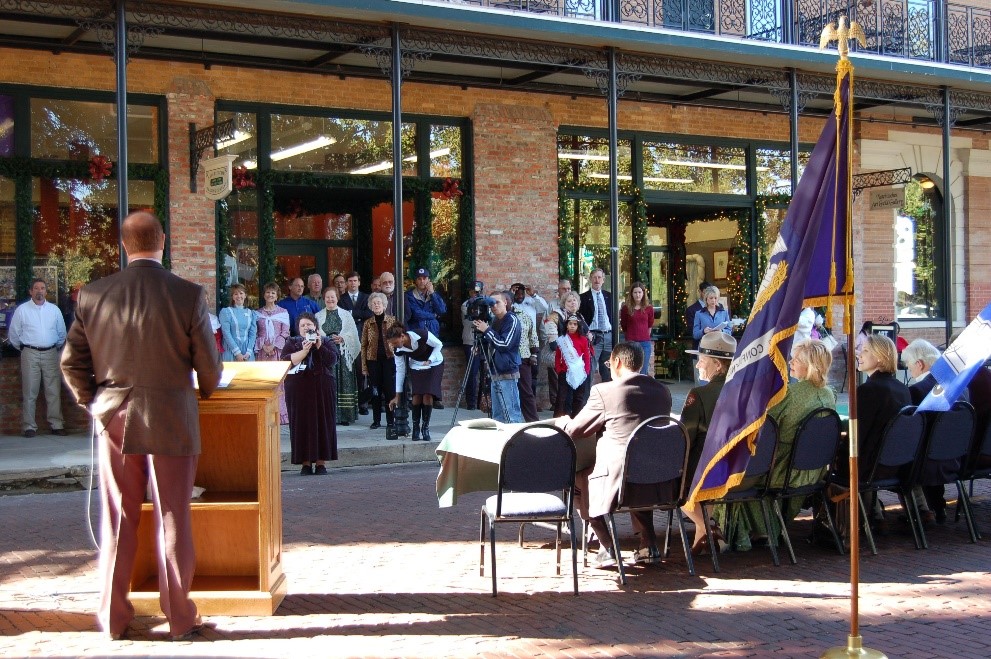 A photograph of a man standing at a podium speaking to a crowd of people standing under an overhang of a building. There is a purple flag and other individuals sitting a table in the foreground of the image.