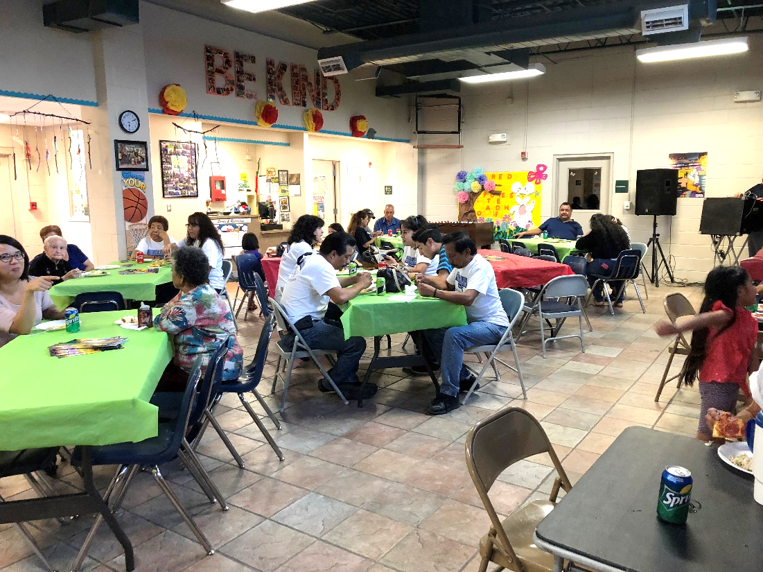 A photograph of a room full of people sitting at tables and eating together at a potluck meal.