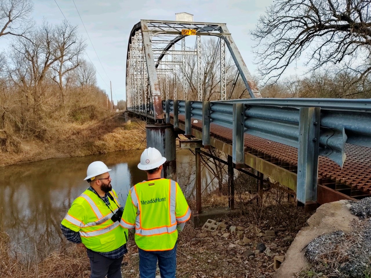 A photograph of two workers looking at the under structure of a bridge that stretches over a river.