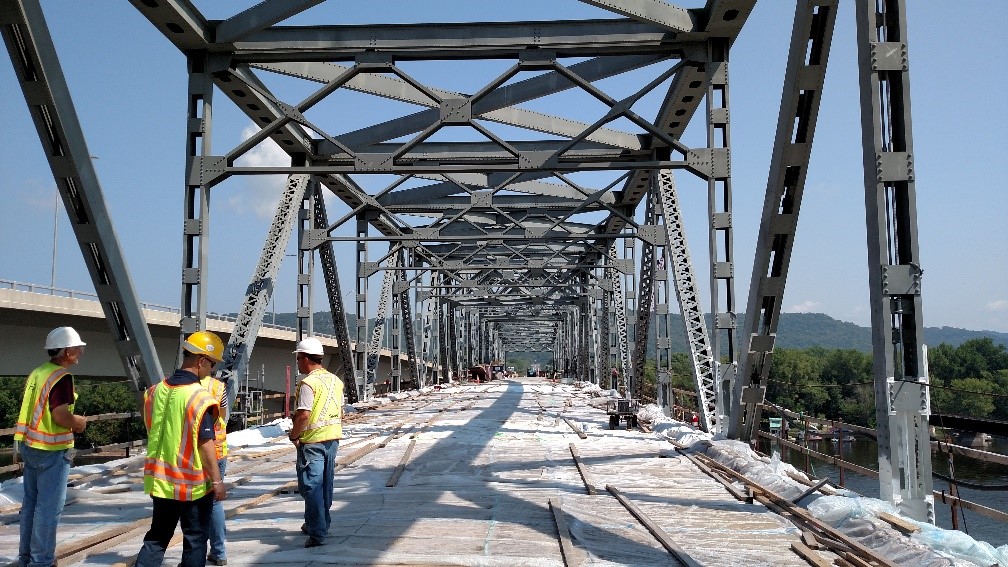 Three men with hard hats and safety vests standing on the partially built truss bridge.