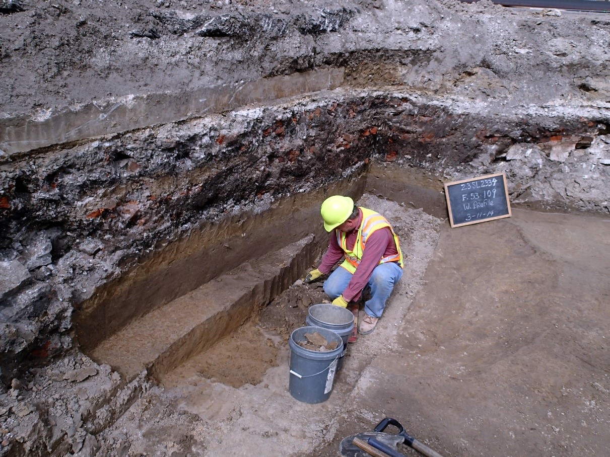 A photograph of a worker in a hard hat and safety vest digging in a archeological site.