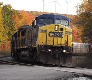 Photograph of a CSX train curving around a bend. The train tracks are lined with dense, colorful fall foilage. Four wind turbines rise beyond and behind the trees.