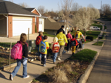 Photograph of a group of elementary school students and their parents walking along a sidewalk in a suburban neighborhood