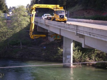 Photograph of a cherry picker truck on top of a bridge. The truck's arm is extended out, around, and under the bridge, where a worker is dislodging nests from the bridge